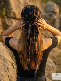 A woman with long hair, seen from behind, stands outdoors touching a black and brown tie-dye headscarf. She wears a black off-shoulder top and is surrounded by large rocks.