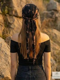 A person with long hair, wearing a black off-shoulder top and dark jeans, stands outdoors facing away. They have a black patterned scarf tied around their head. Rocks and greenery are in the background.