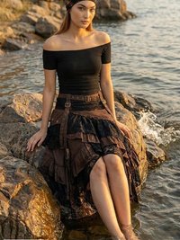 A woman in a black off-shoulder top and layered brown skirt sits on rocks by the water, with her feet in the stream. She wears a dark headscarf and gazes calmly at the camera.