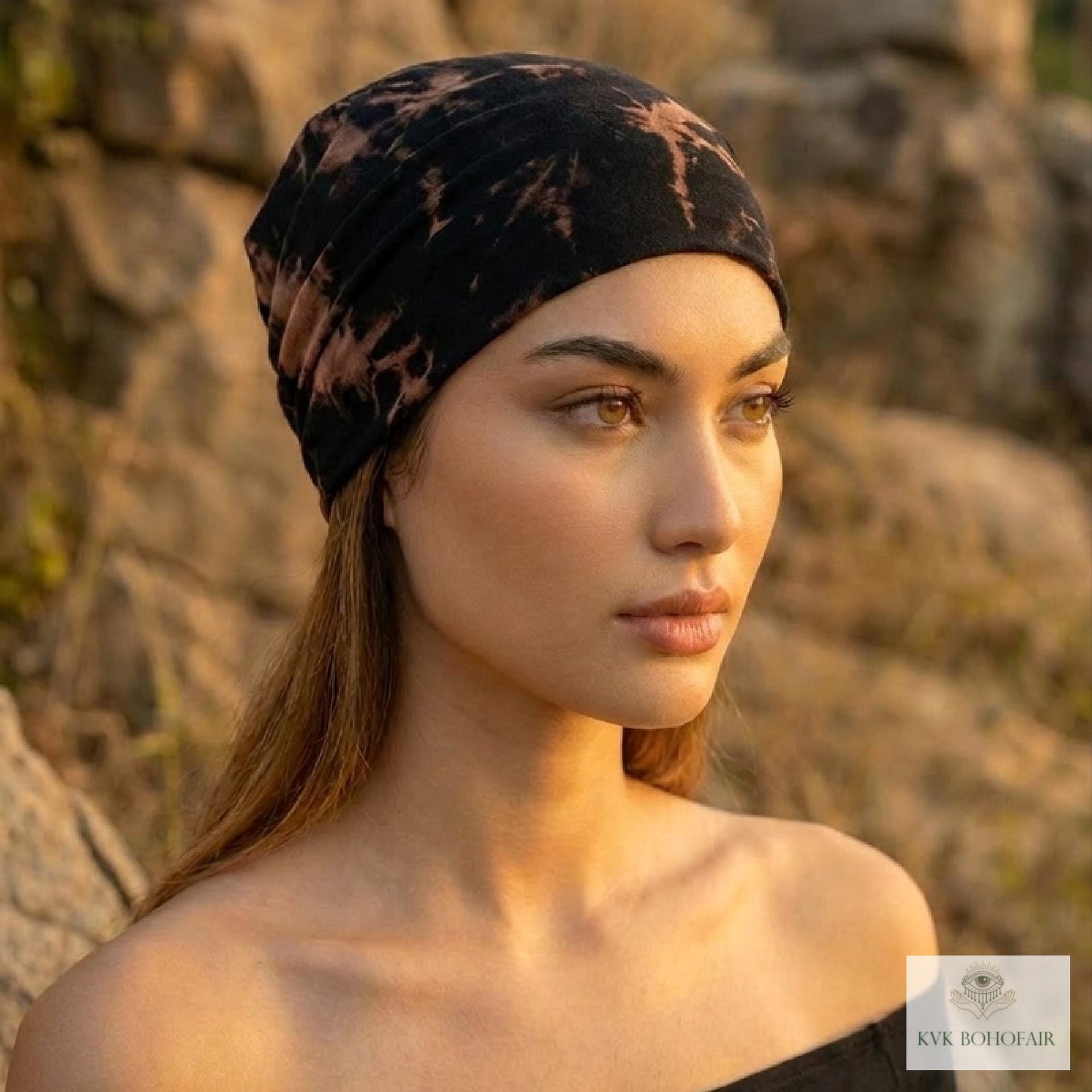 A woman with long hair wearing a black and brown tie-dye headscarf gazes into the distance outdoors, with sunlight illuminating her face. Rocks and warm natural light create a serene, earthy atmosphere.