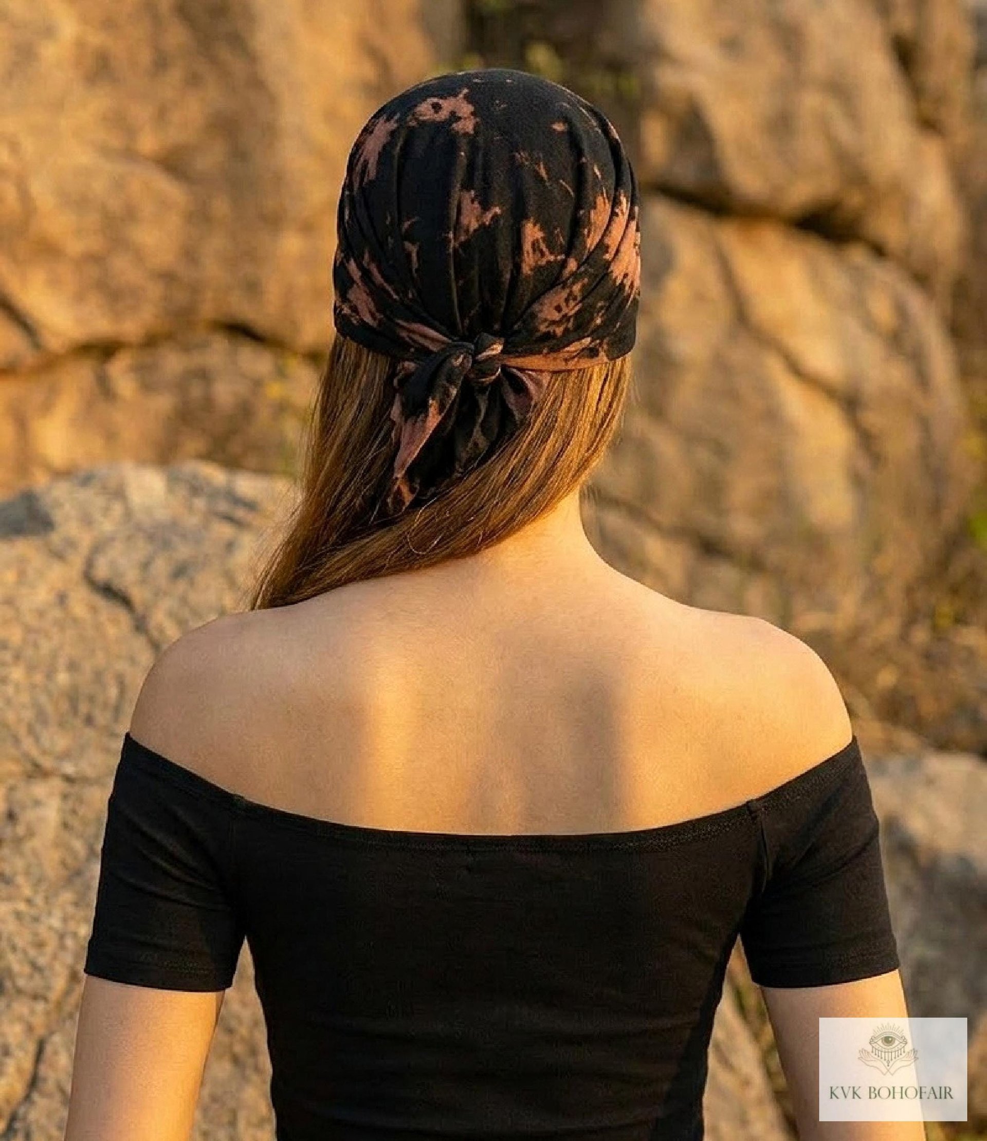 A person with long hair wears an off-shoulder black top and a black and brown headscarf, standing outdoors with rocky terrain in the background.