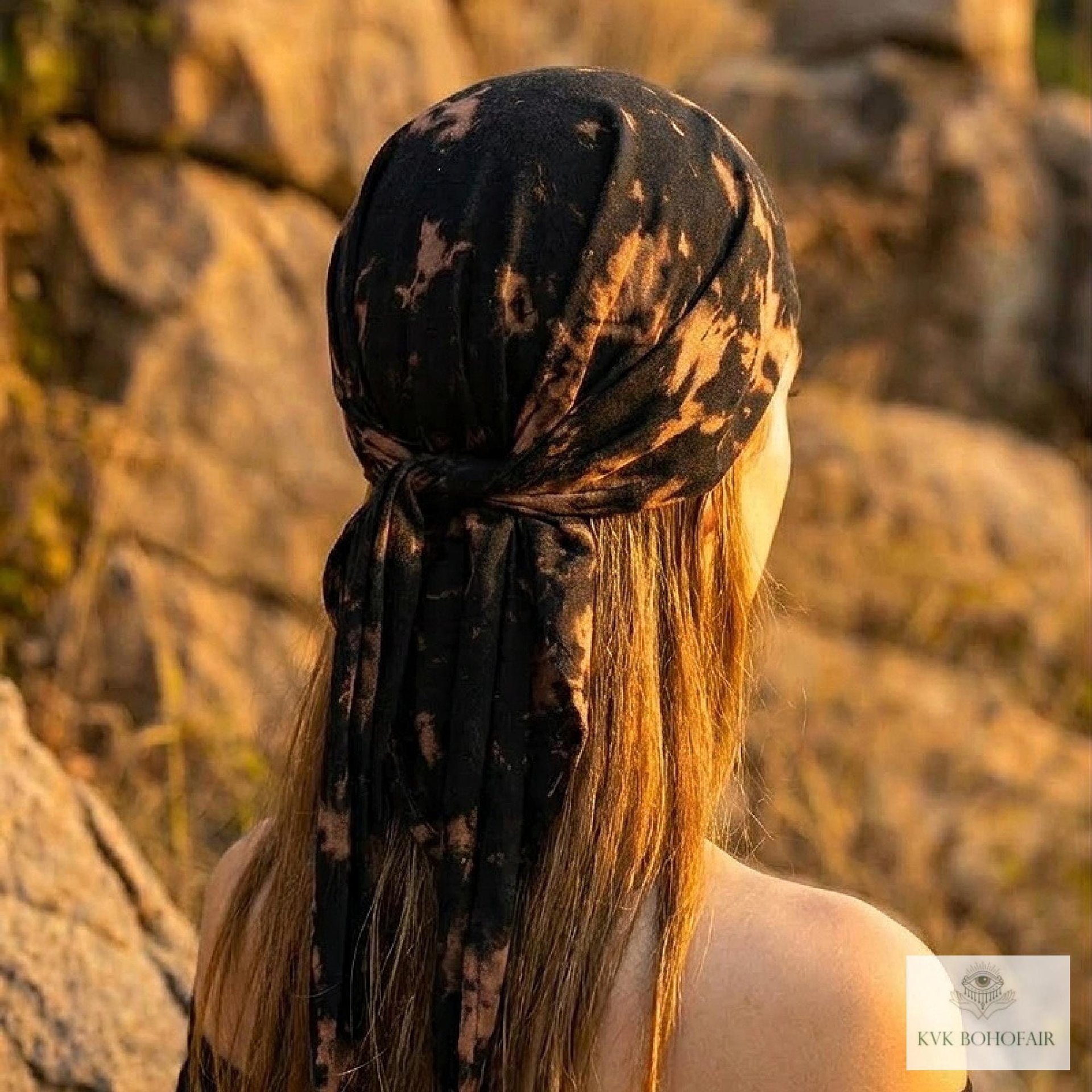 A person with long hair, seen from behind, wears a black and tan tie-dye headscarf outdoors near rocks. Warm sunlight highlights their hair.