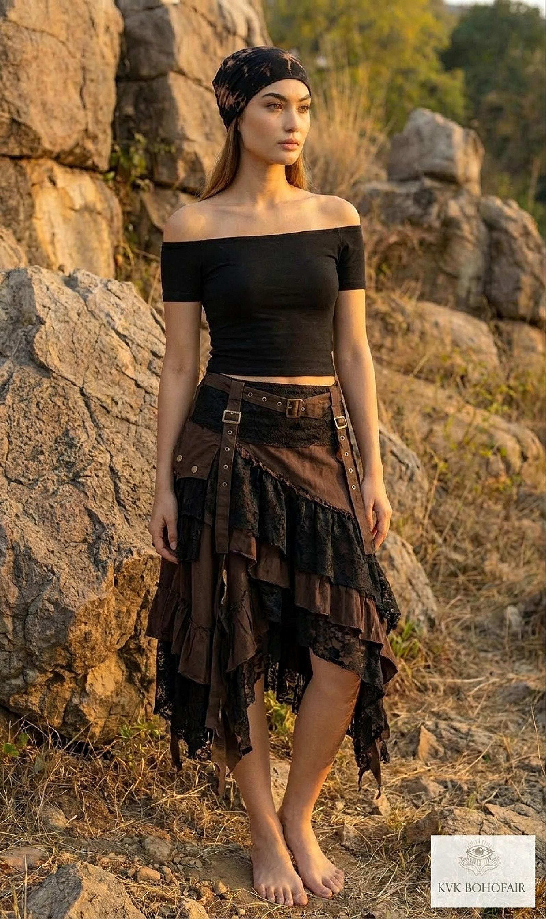 A woman in a black off-shoulder top, layered brown skirt, and headscarf stands barefoot on rocky terrain, surrounded by large rocks and dry grass, with the sun casting a warm light.