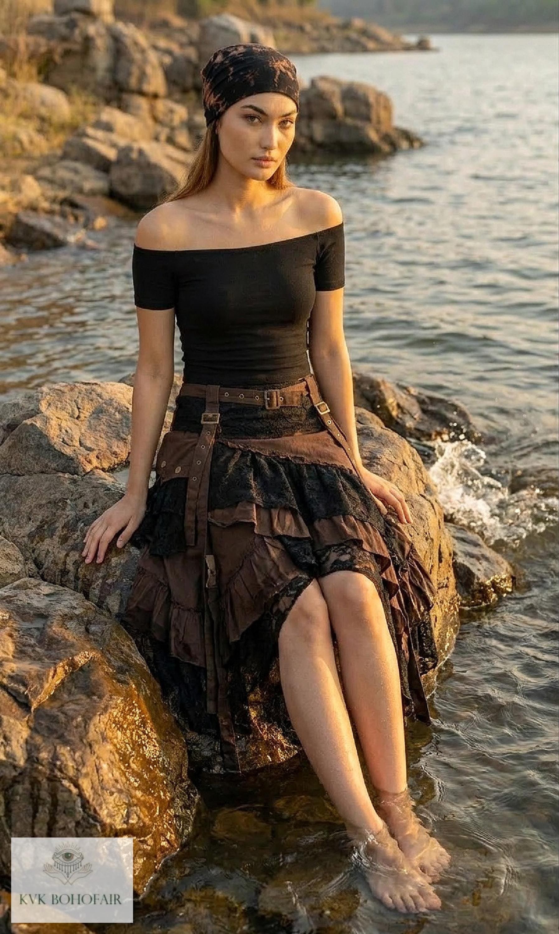A woman in a black off-shoulder top and layered brown skirt sits on rocks by the water, with her feet in the stream. She wears a dark headscarf and gazes calmly at the camera.