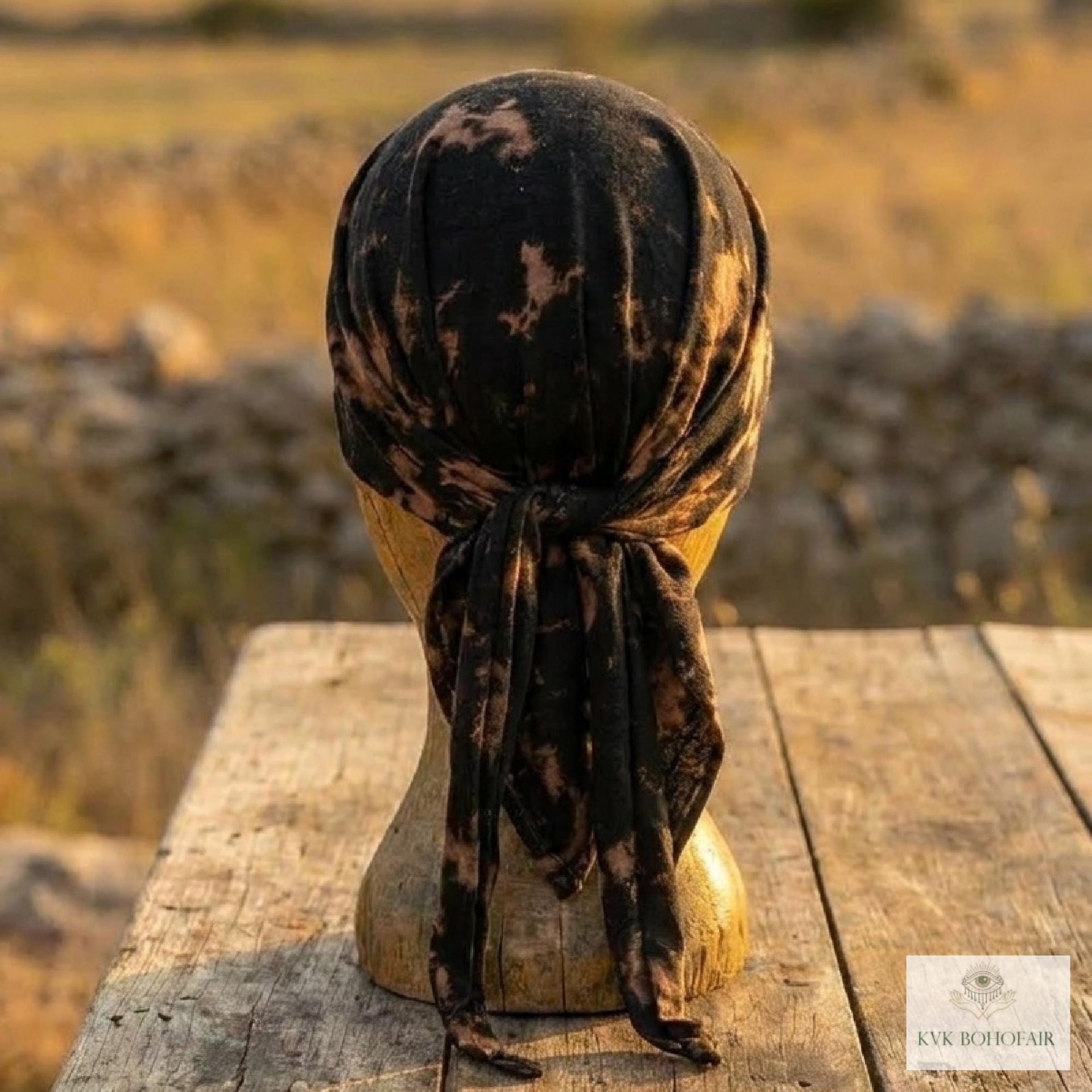 A black and brown tie-dye headscarf is tied on a mannequin head, placed on a rustic wooden table outdoors, with a blurred field and stone wall in the background.