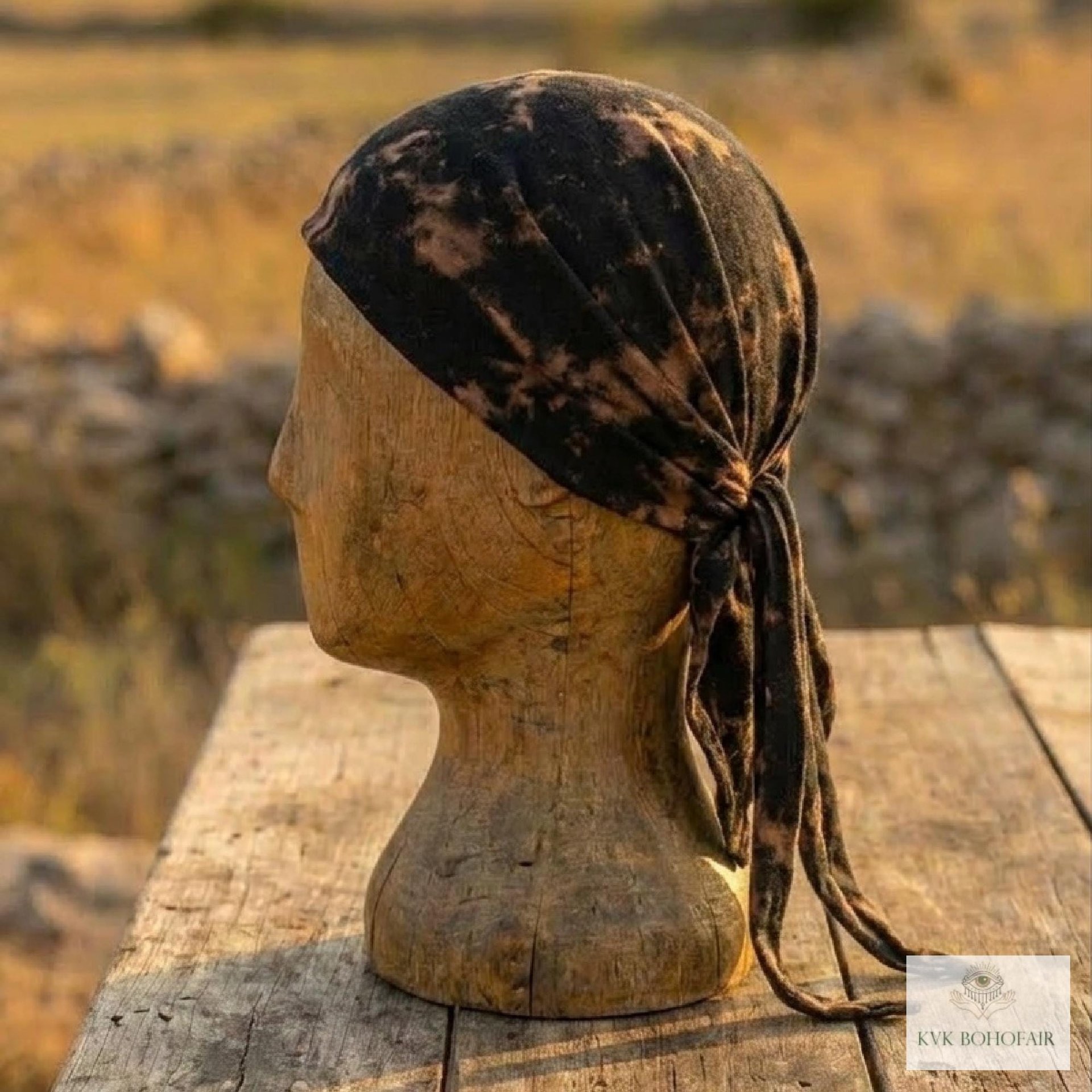 A wooden mannequin head displays a black and tan tie-dye headscarf, tied at the back, set outdoors on a wooden table with a blurred field and stone wall in the background.