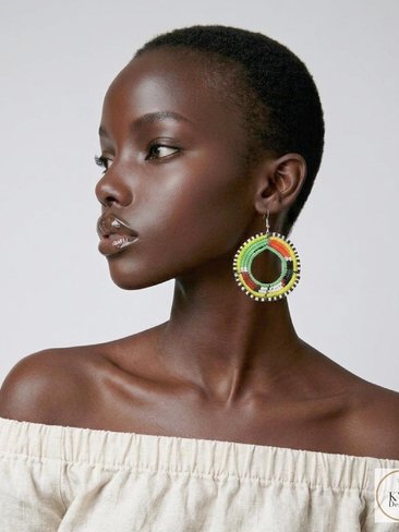A woman with dark skin and short hair wears a cream off-shoulder top and colorful beaded hoop earrings. She looks to the side against a plain background.