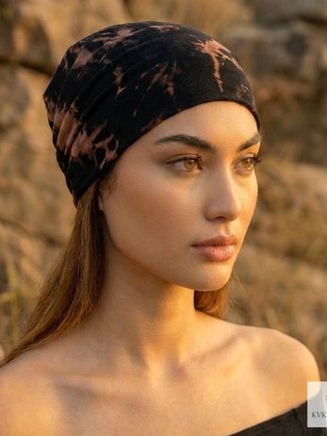 A woman with long hair wearing a black and brown tie-dye headscarf gazes into the distance outdoors, with sunlight illuminating her face. Rocks and warm natural light create a serene, earthy atmosphere.